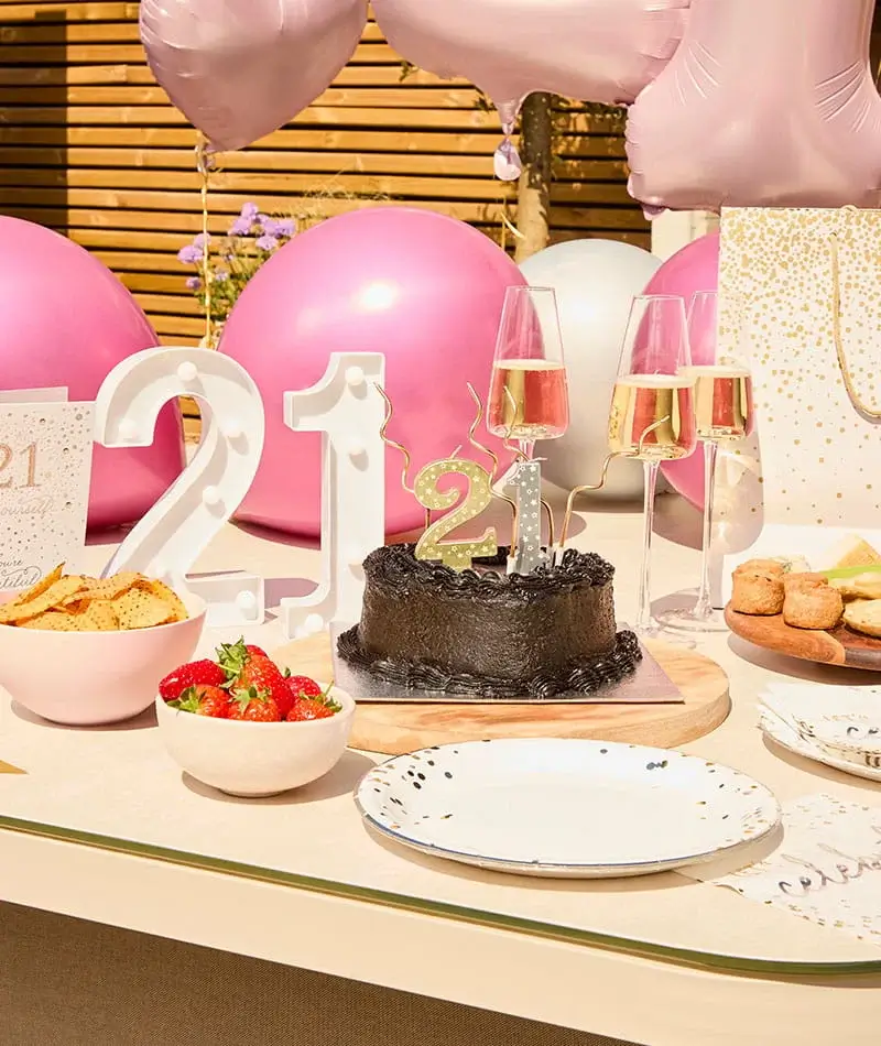 21st birthday celebration table with a chocolate cake, number candles, champagne glasses, pink balloons, snacks, and party decorations.